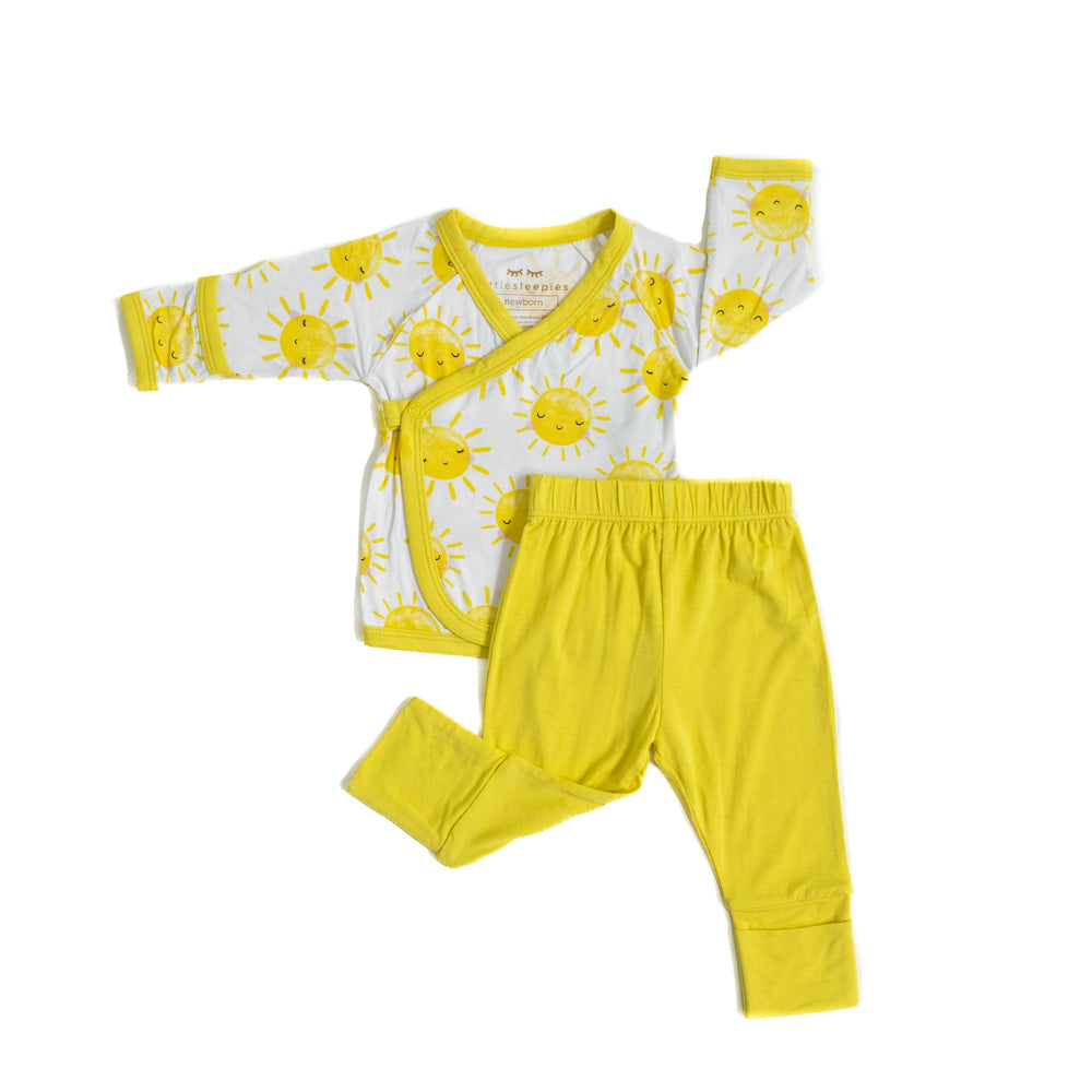 Flat lay image of a two-piece crossover set that features a long sleeve wrap style top with snap closures and coordinating pants with convertible footies. This style pictured has the wrap style top in Sunshine print with sweet yellow sunshines on a white brown background with matching yellow trim.