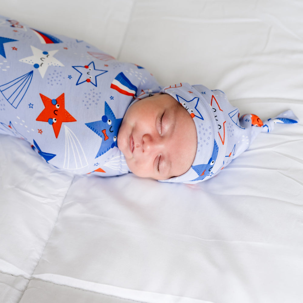 Close up image of a child laying on a bed swaddled in a Blue Stars and Stripes printed swaddle and hat set