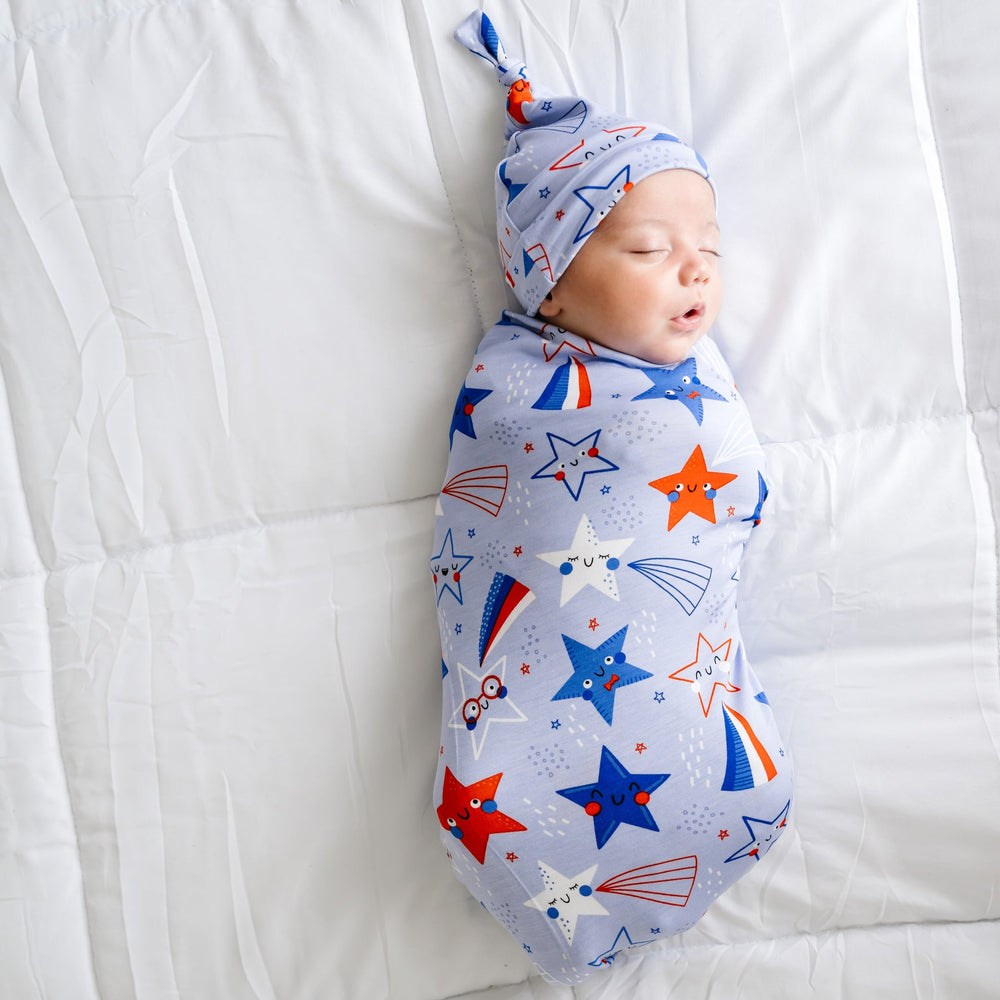 Child laying on a bed swaddled in a Blue Stars and Stripes printed swaddle and hat set