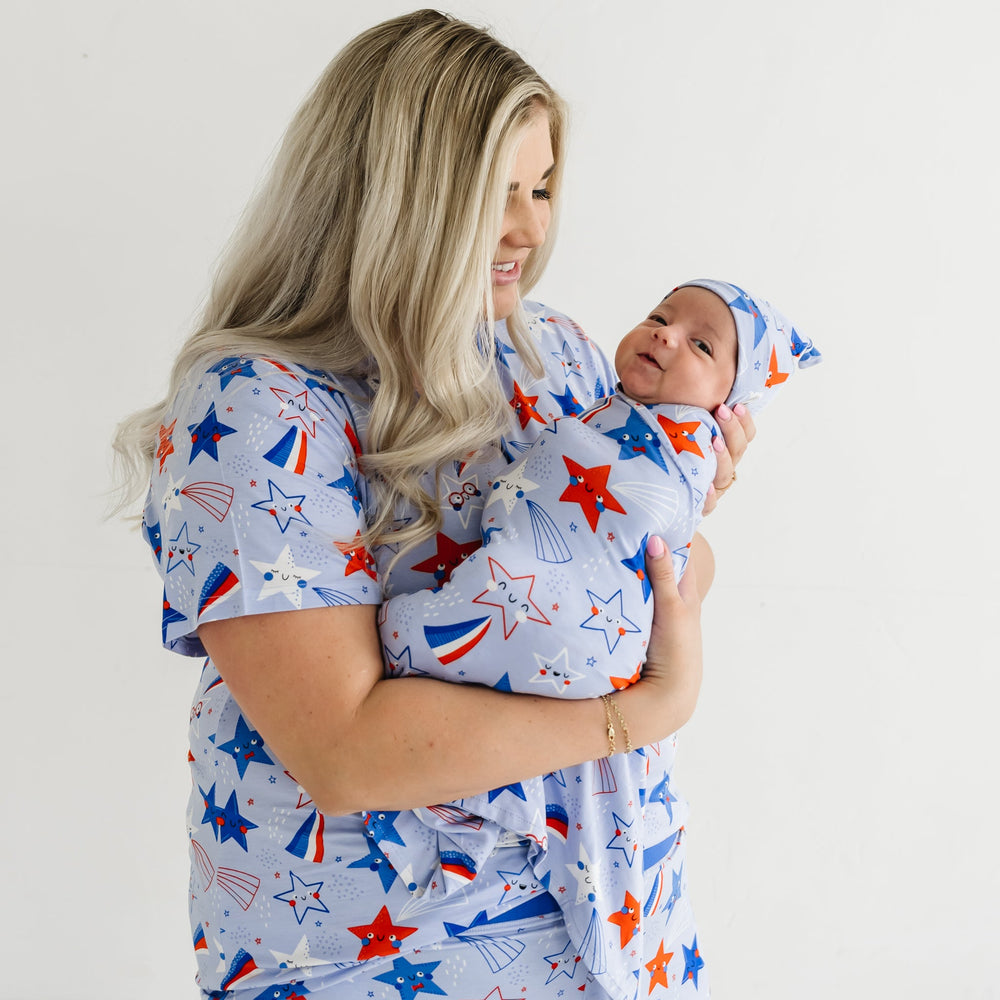 Mother wearing a women's Blue Stars and Stripes printed pajama top while holding her newborn swaddled in a Blue Stars and Stripes printed swaddle and hat set