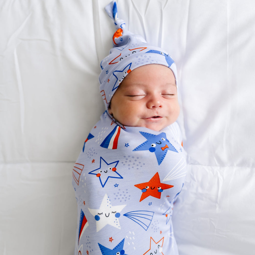 Child laying on a bed swaddled in a Blue Stars and Stripes printed swaddle and hat set