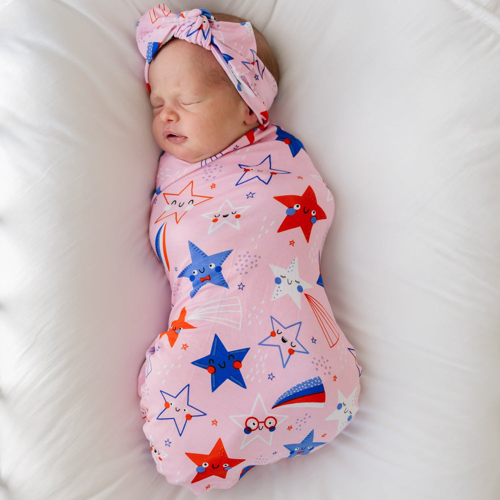 Child laying on a bed swaddled in a Pink Stars and Stripes printed swaddle and headband set