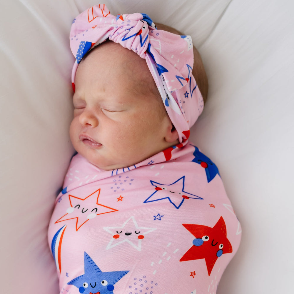Close up image of a child laying on a bed swaddled in a Pink Stars and Stripes printed swaddle and headband set