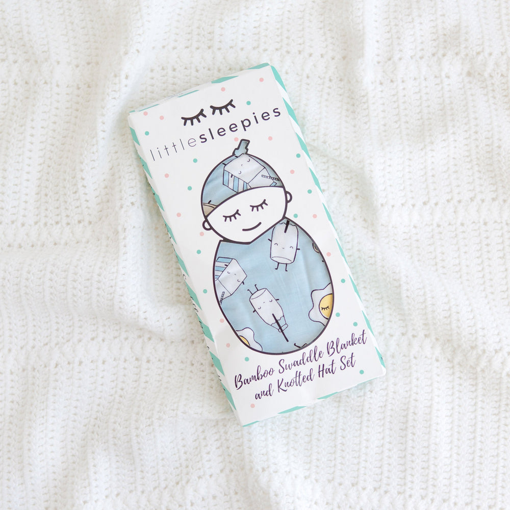 Image of Blue Breakfast Buddies printed swaddle and hat set in Little Sleepies packaging. This print has a light blue background with white trim accents and the breakfast foods featured on this print include sunny side up eggs, toast, and milk.