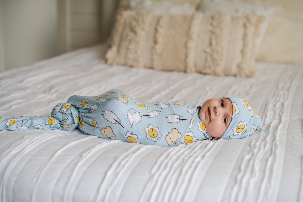 Image of baby boy in Blue Breakfast Buddies printed swaddle and hat set. This print has a light blue background and the breakfast foods featured on this print include sunny side up eggs, toast, and milk.