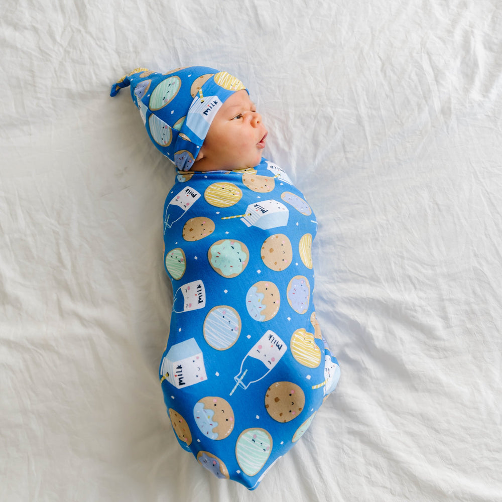 Click to see full screen - Image of infant baby wearing a swaddle and hat set in cookies and milk print. This print features milk cartons, colorful sprinkled cookies, and chocolate chip cookies that sit upon a blue background.