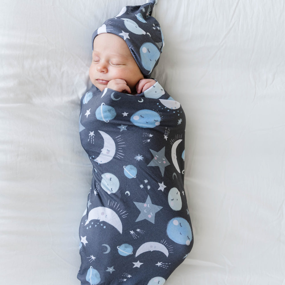 Click to see full screen - Image of infant boy in a Blue To the Moon & Back printed swaddle and hat set. This print features blue and gray moons, stars, and planets on a charcoal background
