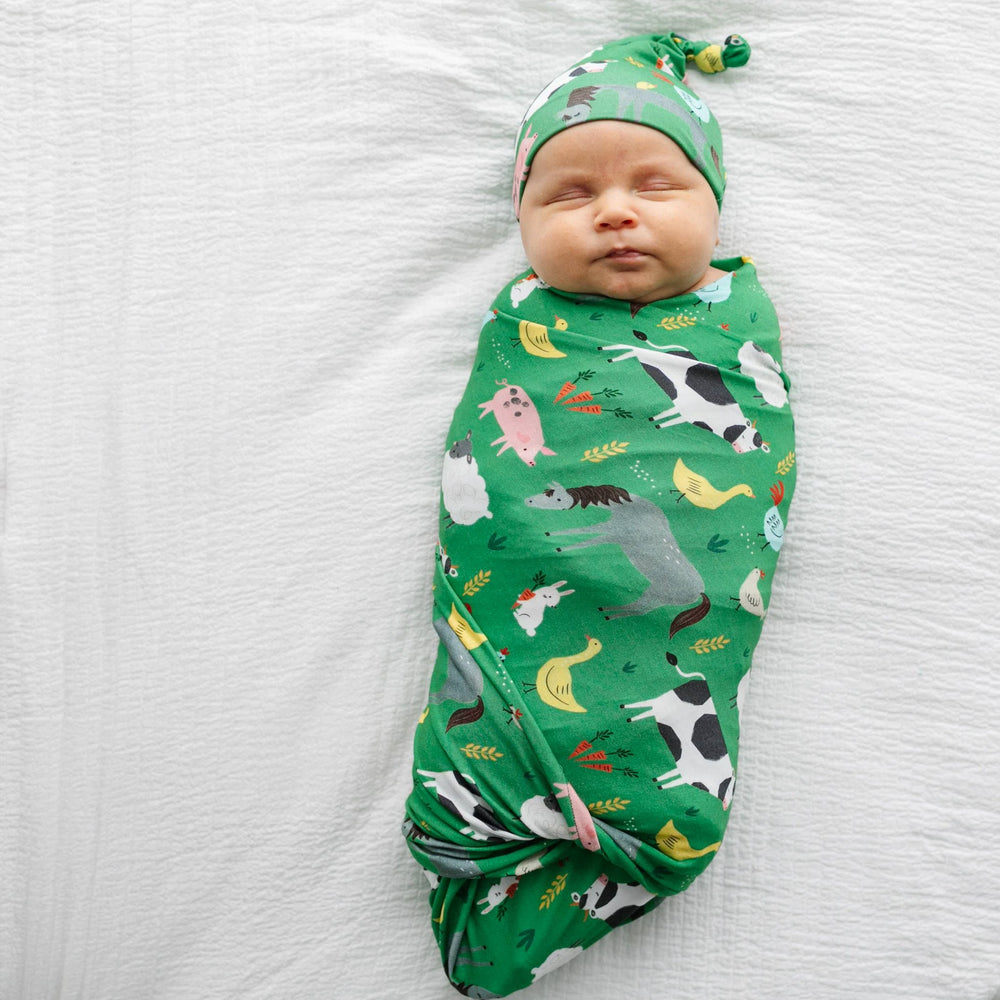 Image of infant boy wearing a swaddle and hat set in green farm animals print. This print includes a green background, and features farm animals such as cows, pigs, ducks, sheep, pigs, chickens, and bunnies.