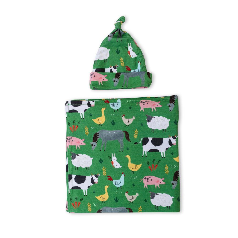 Flat lay image of swaddle and hat set in green farm animals print. This print includes a green background, and features farm animals such as cows, pigs, ducks, sheep, pigs, chickens, and bunnies. 
