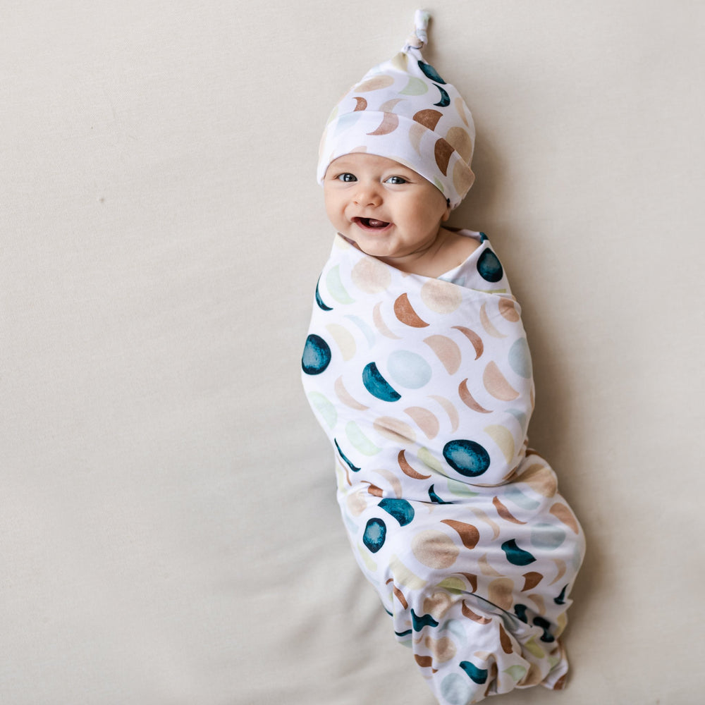 Image of an infant in a swaddle and knotted hat set in Luna Neutral print. This print features phases of the moon in the sweetest shades of creams, tans, and navy watercolor in an all over repeat pattern.