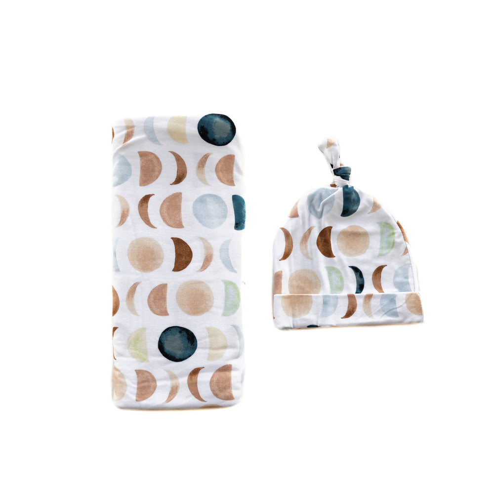 Flat lay image of a swaddle and knotted hat set in Luna Neutral print. This print features phases of the moon in the sweetest shades of creams, tans, and navy watercolor in an all over repeat pattern.