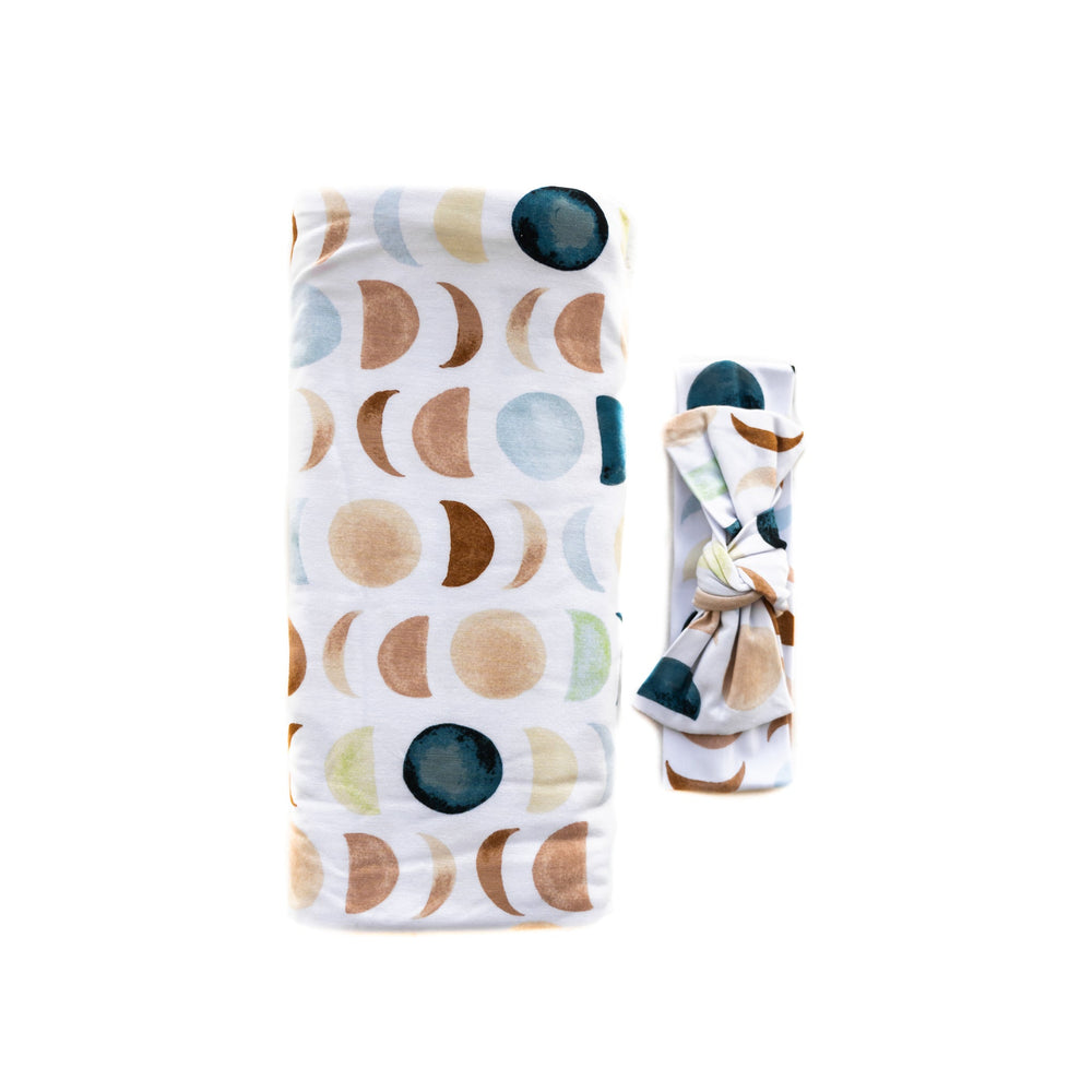 Flat lay image of a swaddle and bow headband set in Luna Neutral print. This print features phases of the moon in the sweetest shades of creams, tans, and navy watercolor in an all over repeat pattern.