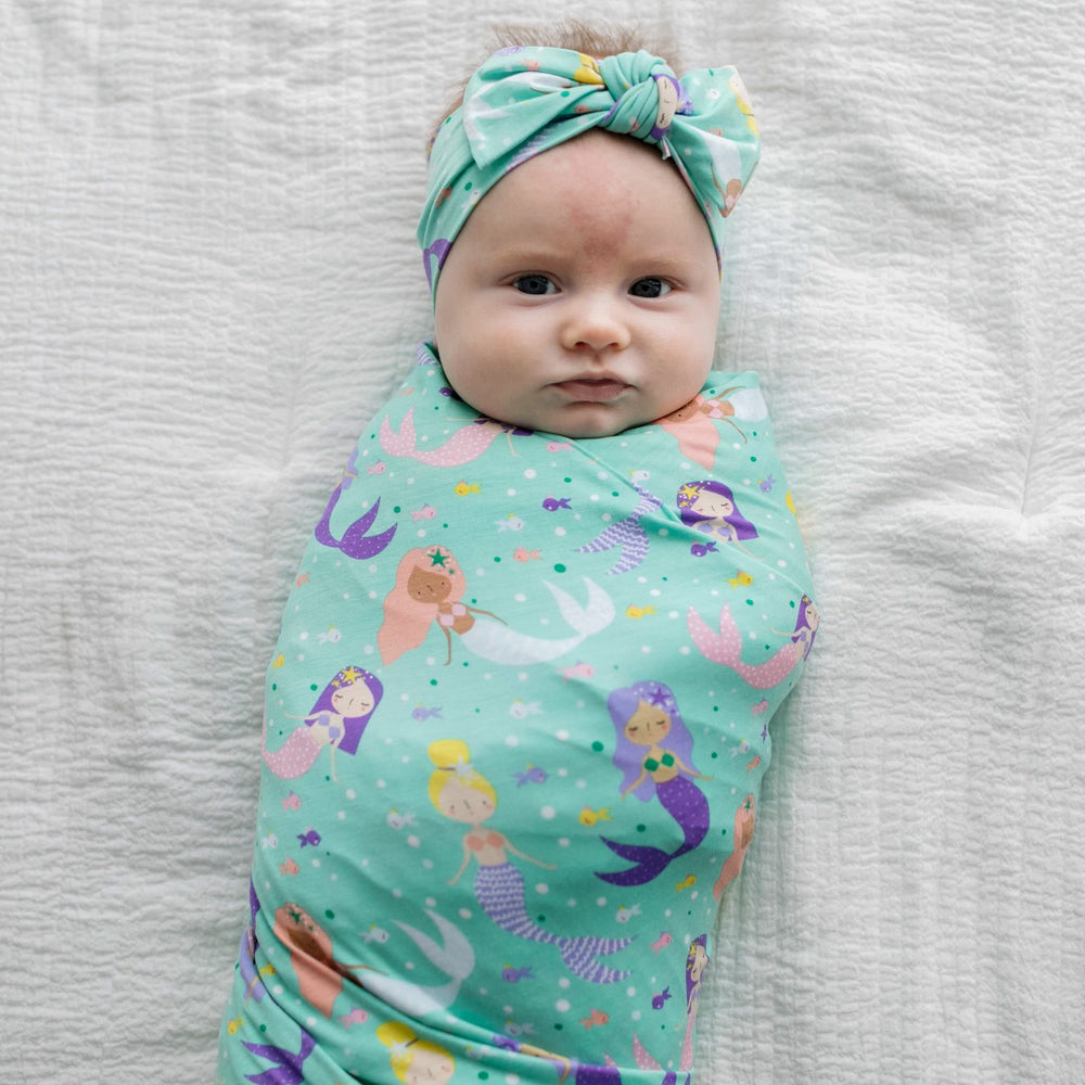 Image of infant girl wearing swaddle and headband set in mermaid print. This print includes multi-colored mermaids and fish that are featured on an aqua background.