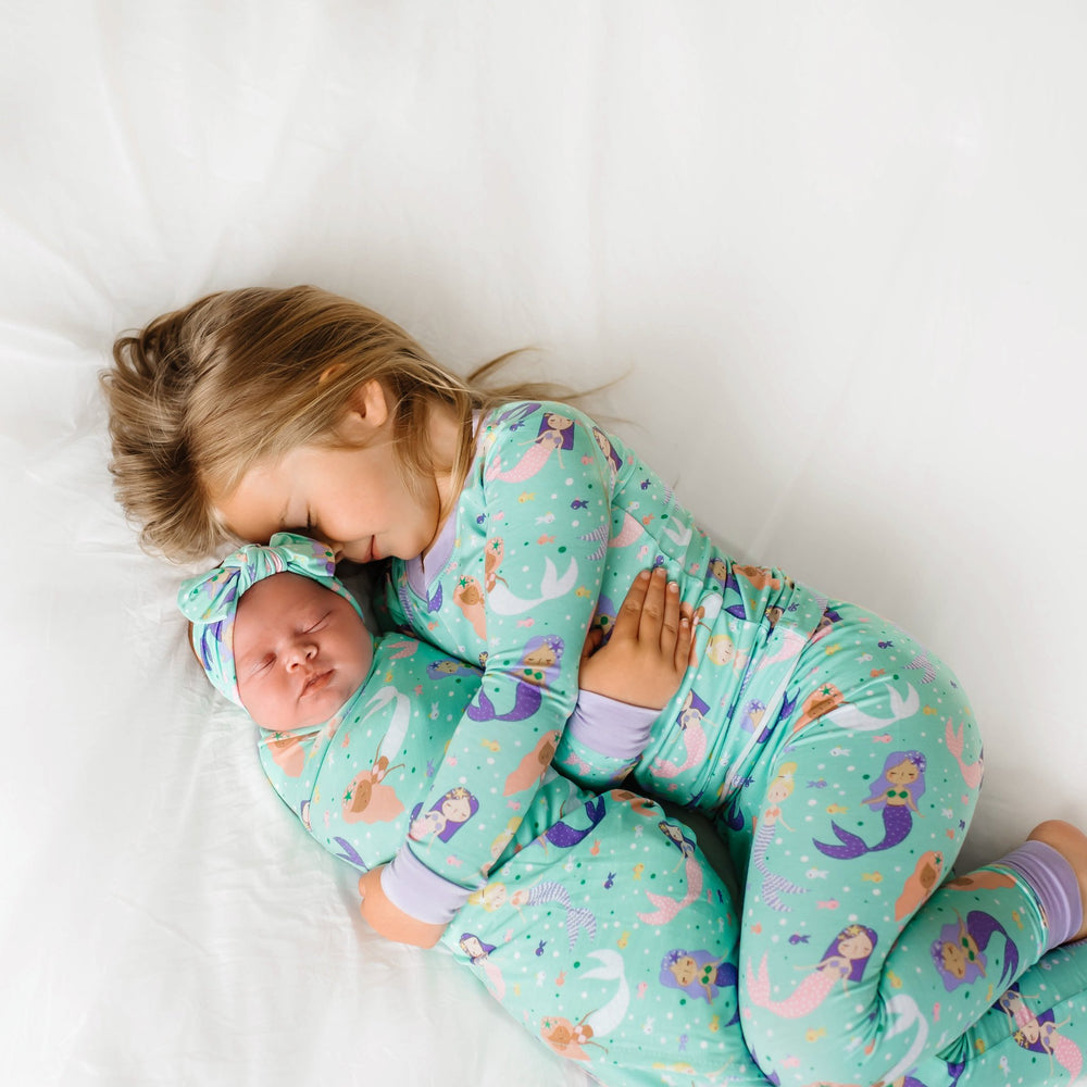 Image of little girl hugging an infant girl. They are both shown wearing matching mermaid print, with the little girl in a two-piece pajama set and the infant girl in a swaddle and headband set. This print includes multi-colored mermaids and fish that are