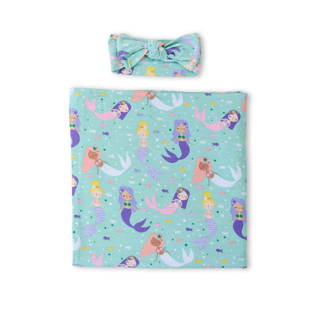 Flat lay image of swaddle and headband set in mermaid print. This print includes multi-colored mermaids and fish that are featured on an aqua background 