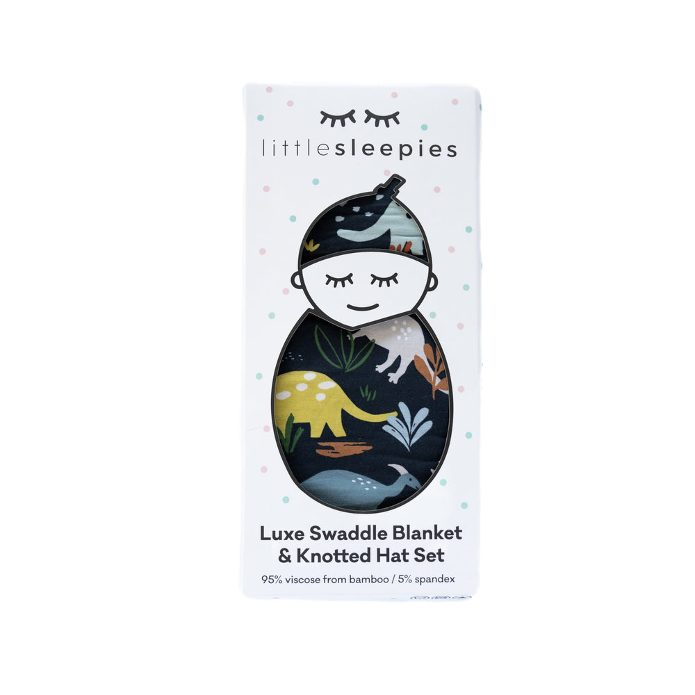 Flat lay image of the packaged peekaboo box of a luxe swaddle blanket and knotted hat set in Navy Jurassic Jungle print. This print features all their prehistoric favorites of various dinosaurs in a bold navy blue to inspire adventurous dreams.