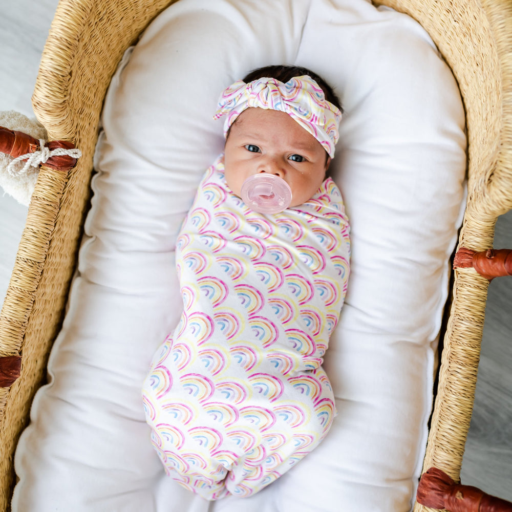 Image of infant girl wearing rainbow printed swaddle and headband set.