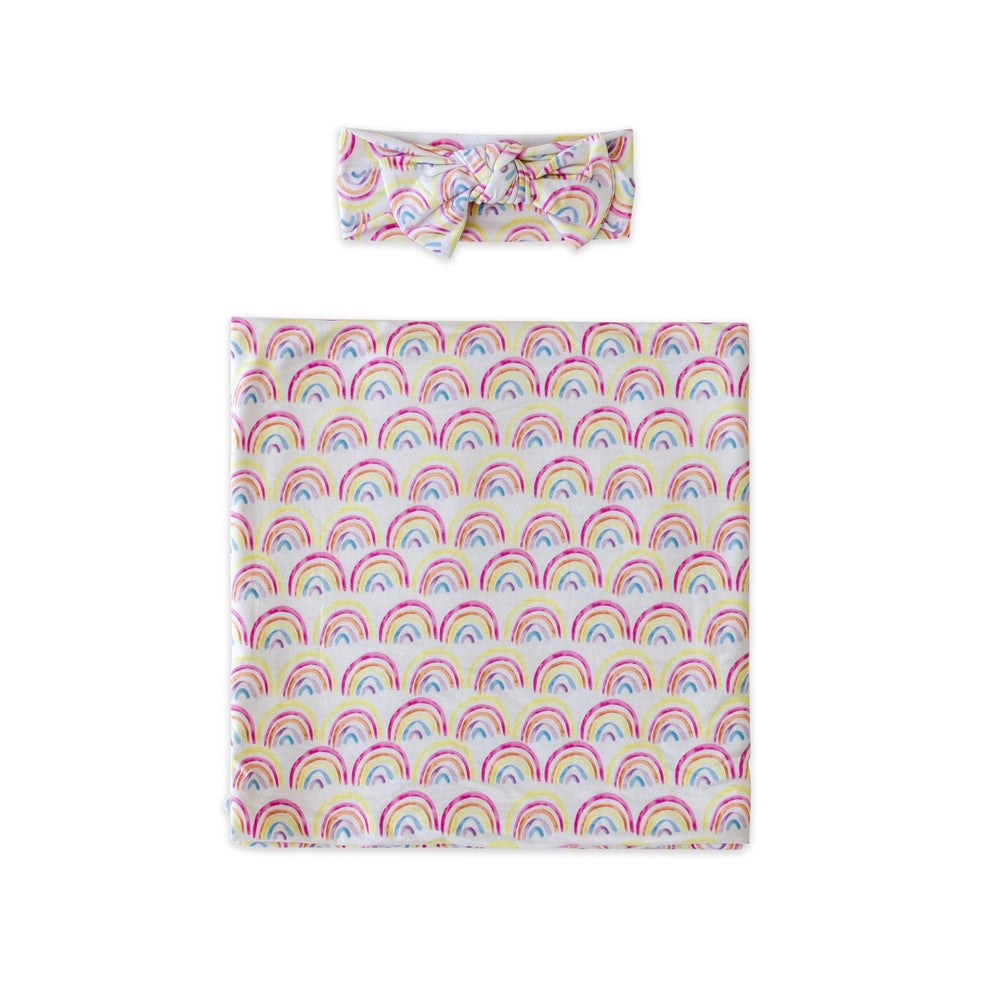 Flat lay image of swaddle and headband set in rainbow print. This print features multicolored rainbows that sit upon a white background