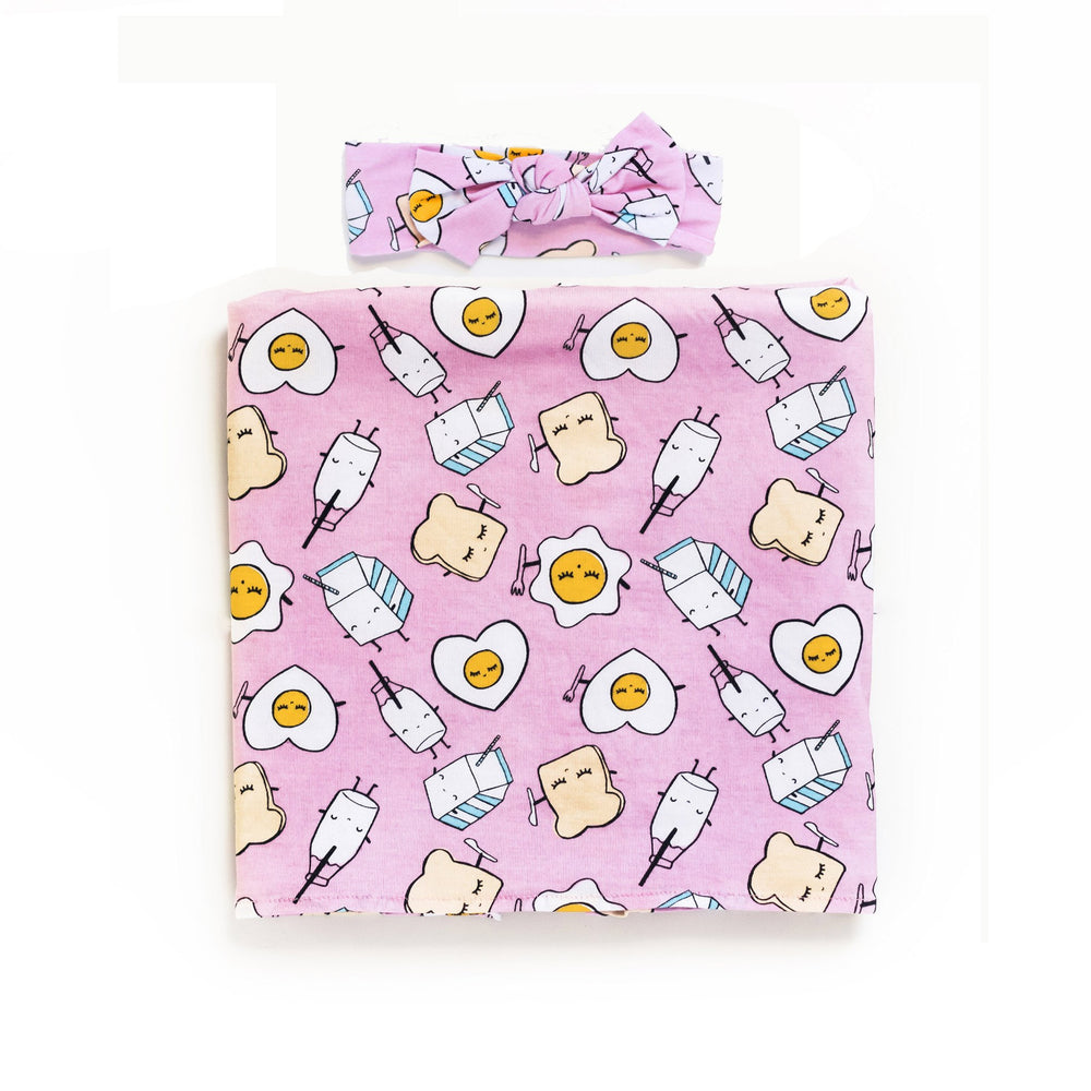 Flat lay image of swaddle and headband set in Pink Breakfast Buddies print. his print has a light pink background and the breakfast foods featured on this print include sunny side up eggs, toast, and milk.