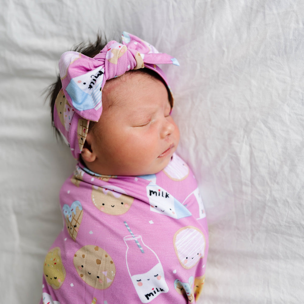Image of infant girl wearing a swaddle and headband set in cookies and milk print. This print features milk cartons, colorful sprinkled cookies, and chocolate chip cookies that sit upon a pink background.