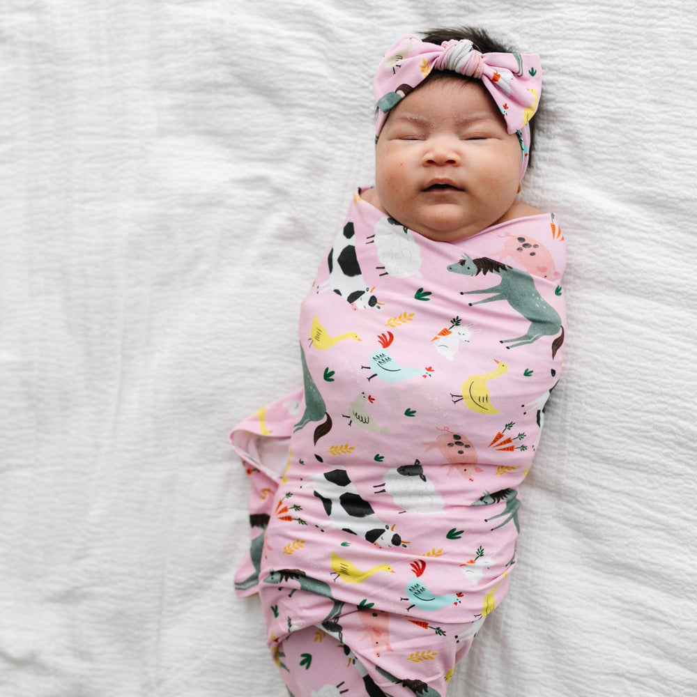 Image of infant girl wearing a swaddle and headband set in pink farm animals print. This print includes a pink background, and features  farm animals such as cows, pigs, ducks, sheep, pigs, chickens, and bunnies.
