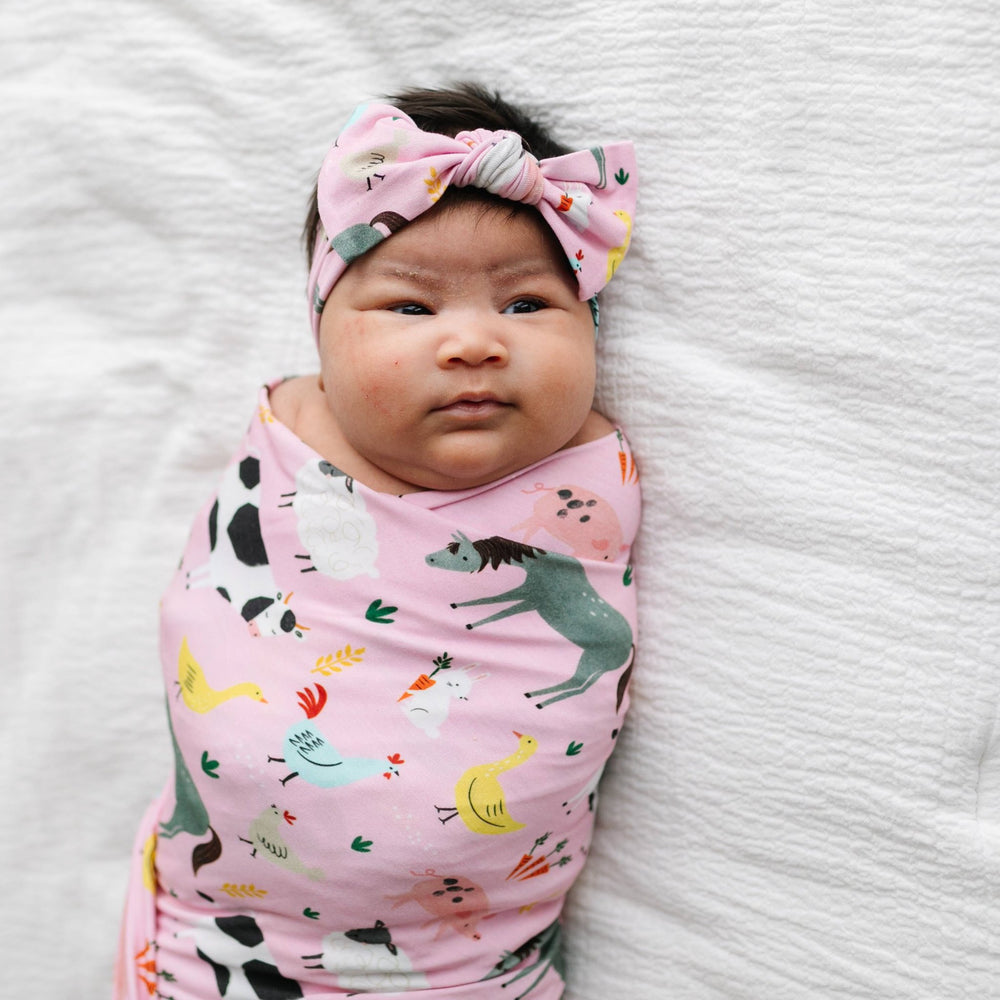Image of infant girl wearing a swaddle and headband set in pink farm animals print. This print includes a pink background, and features  farm animals such as cows, pigs, ducks, sheep, pigs, chickens, and bunnies.