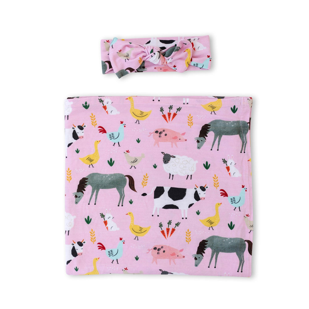 Flat lay image of swaddle and headband set in pink farm animals print. This print includes a pink background, and features farm animals such as cows, pigs, ducks, sheep, pigs, chickens, and bunnies. 