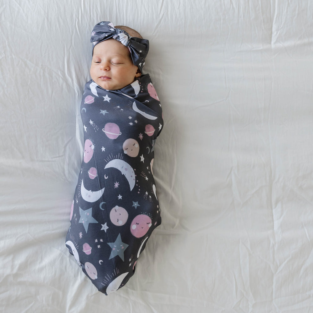 Image of infant girl wearing a swaddle and headband set in pink to the moon and back print. This print features pink and gray moons, stars, and planets on a charcoal background