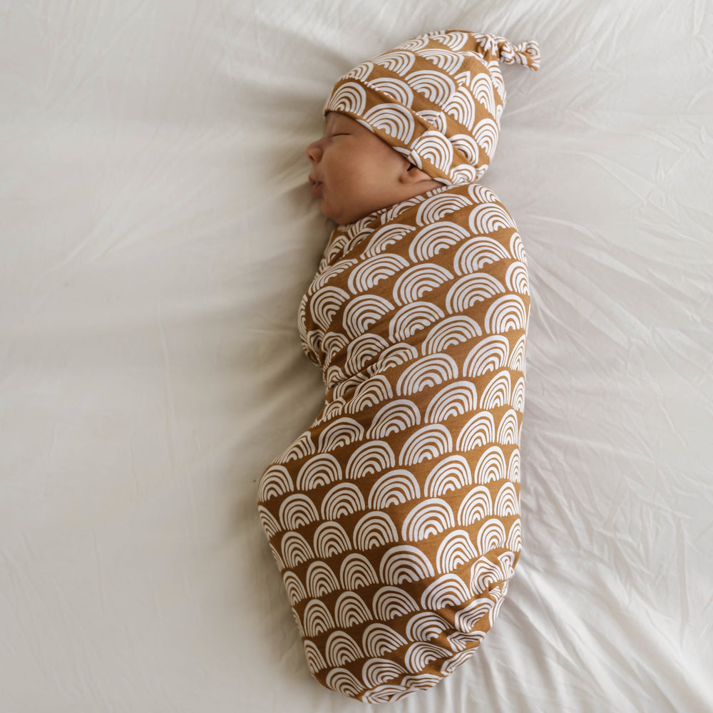 Image of infant boy wearing a swaddle and hat set in Rust Rainbows print. This print features white rainbows that sit upon a rust brown background.