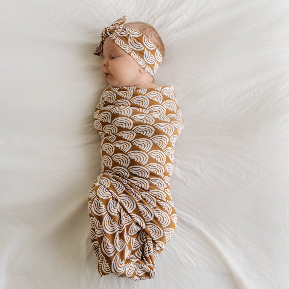 Image of infant girl wearing a swaddle and headband set in Rust Rainbows print. This print features white rainbows that sit upon a rust brown background
