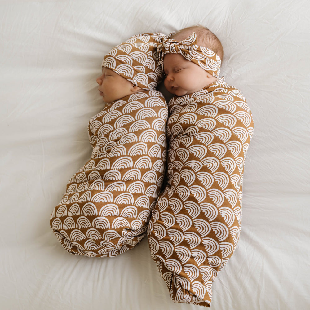 Image of infant boy and girl wearing matching swaddle sets in Rust Rainbows print. This print features white rainbows that sit upon a rust brown background