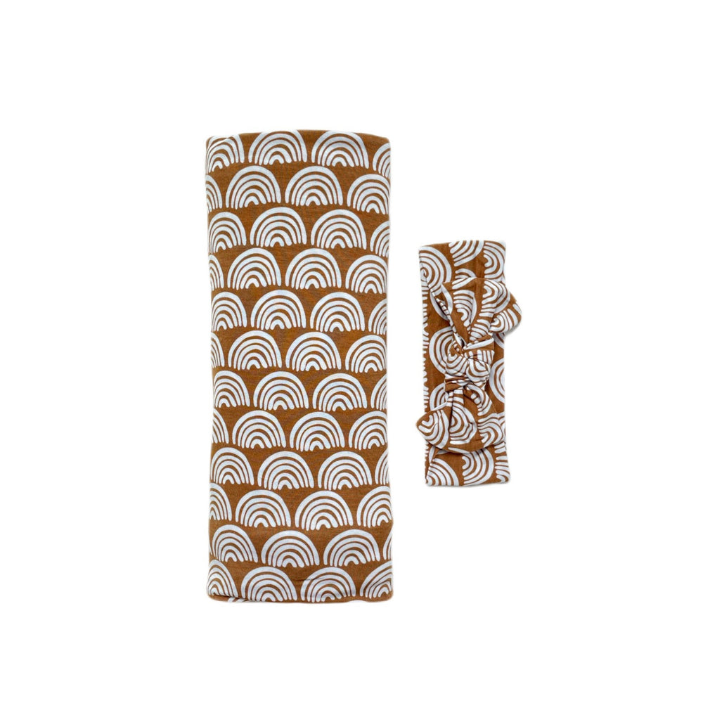 Click to see full screen - Flat lay image of swaddle and headband set in Rust Rainbows print. This print features white rainbows that sit upon a rust brown background