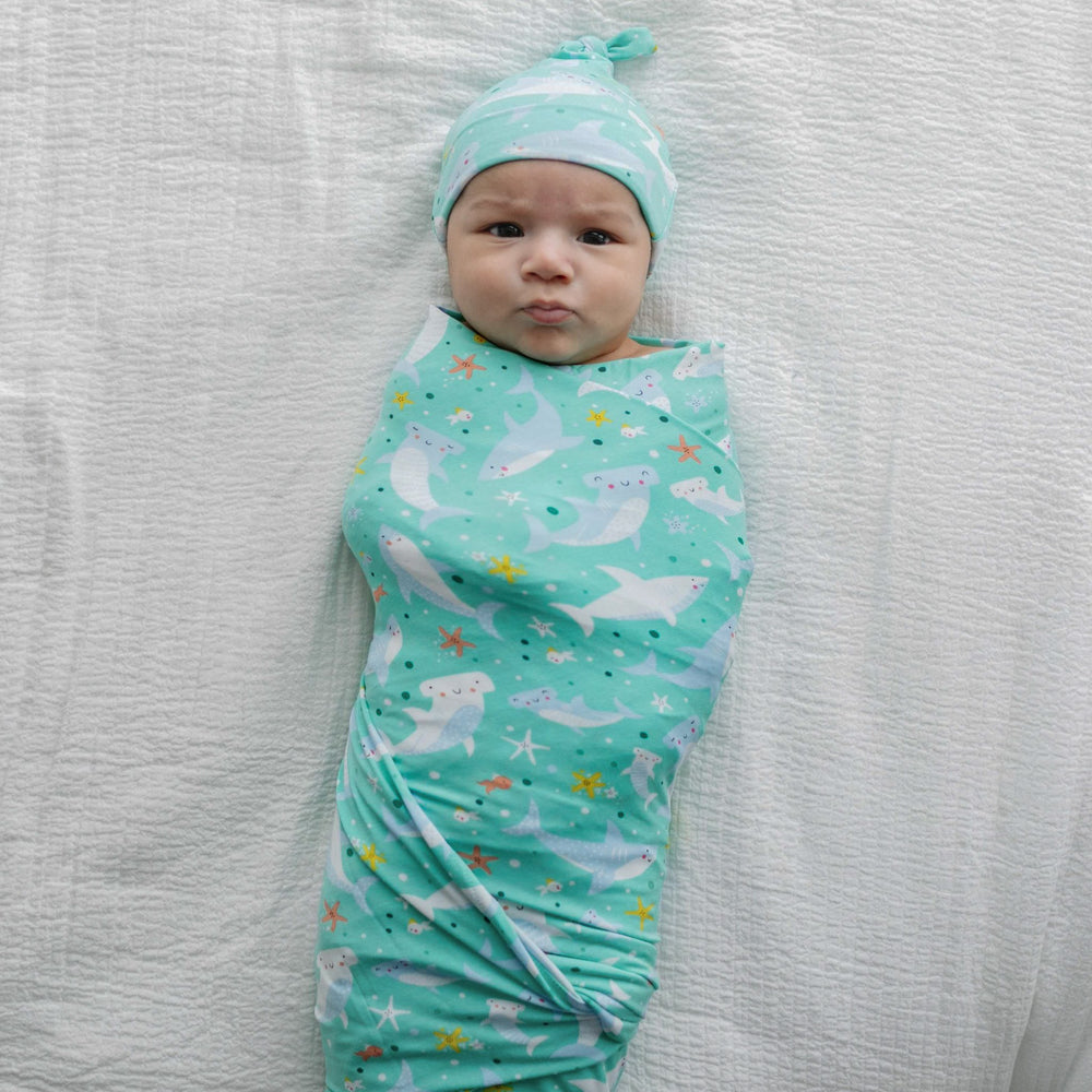 Image of infant boy wearing shark printed swaddle and hat set. This print includes hammerhead and great white sharks, featured on an aqua background