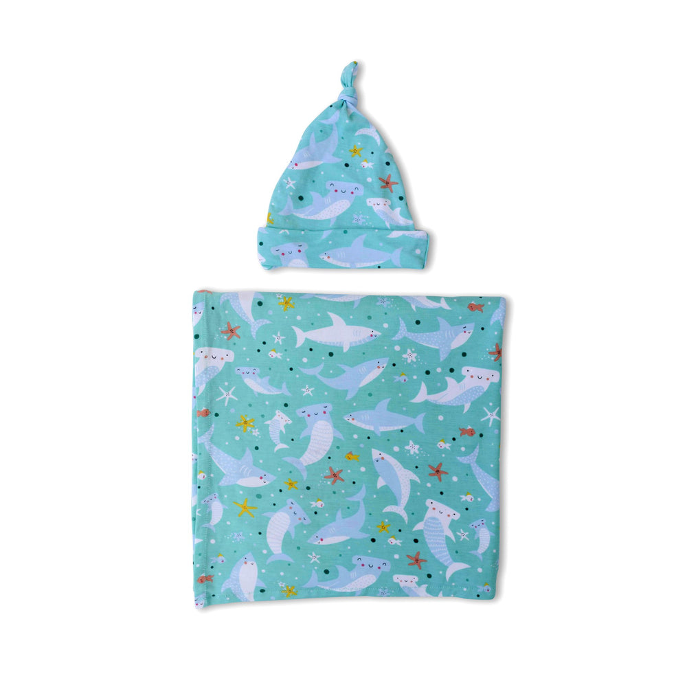 Flat lay image of swaddle and hat set in shark print. This print includes hammerhead and great white sharks, featured on an aqua background