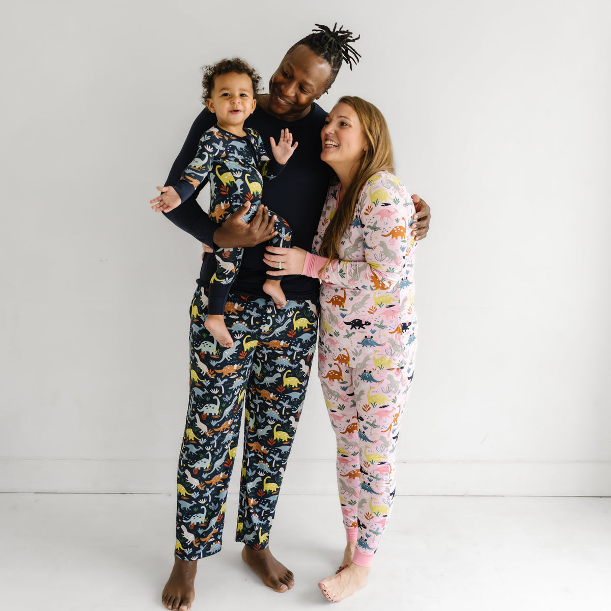 Family of three together wearing coordinating Jurassic Jungle printed pajamas. Father and son are matching wearing Navy Jurassic Jungle prints in men's pajama bottoms, and zippy styles. Mom is wearing Pink Jurassic Jungle in women's pants and top.  