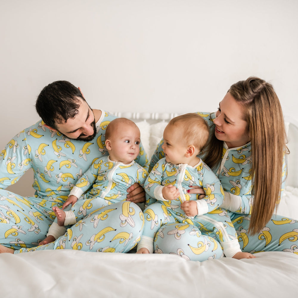 Family of four all wearing matching banana printed pajamas. The mom and dad pajama sets have a light blue background with white trim accents and pops of yellow coming from the banana print. While the two baby boy's zip up rompers have a light blue background with white trim accents and pops of yellow coming from the banana print