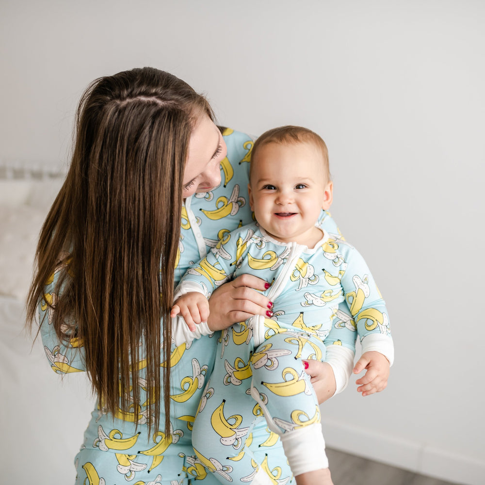 Mother and son both wearing banana printed pajamas. This print features a light blue background with pops of yellow and white trim accents on the collar, sleeve cuffs, and pant cuffs. 