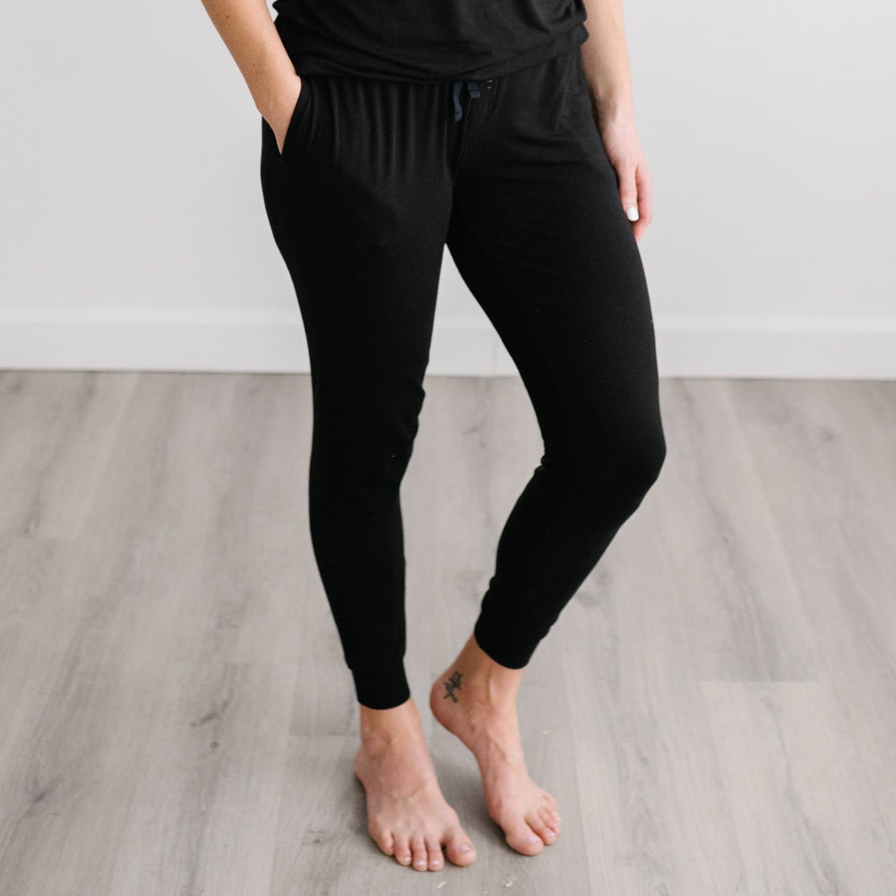 Image of female model wearing solid black pajama pants. The pajama pants feature an adjustable waist drawstring, cuffs at the ankles, and pockets.
