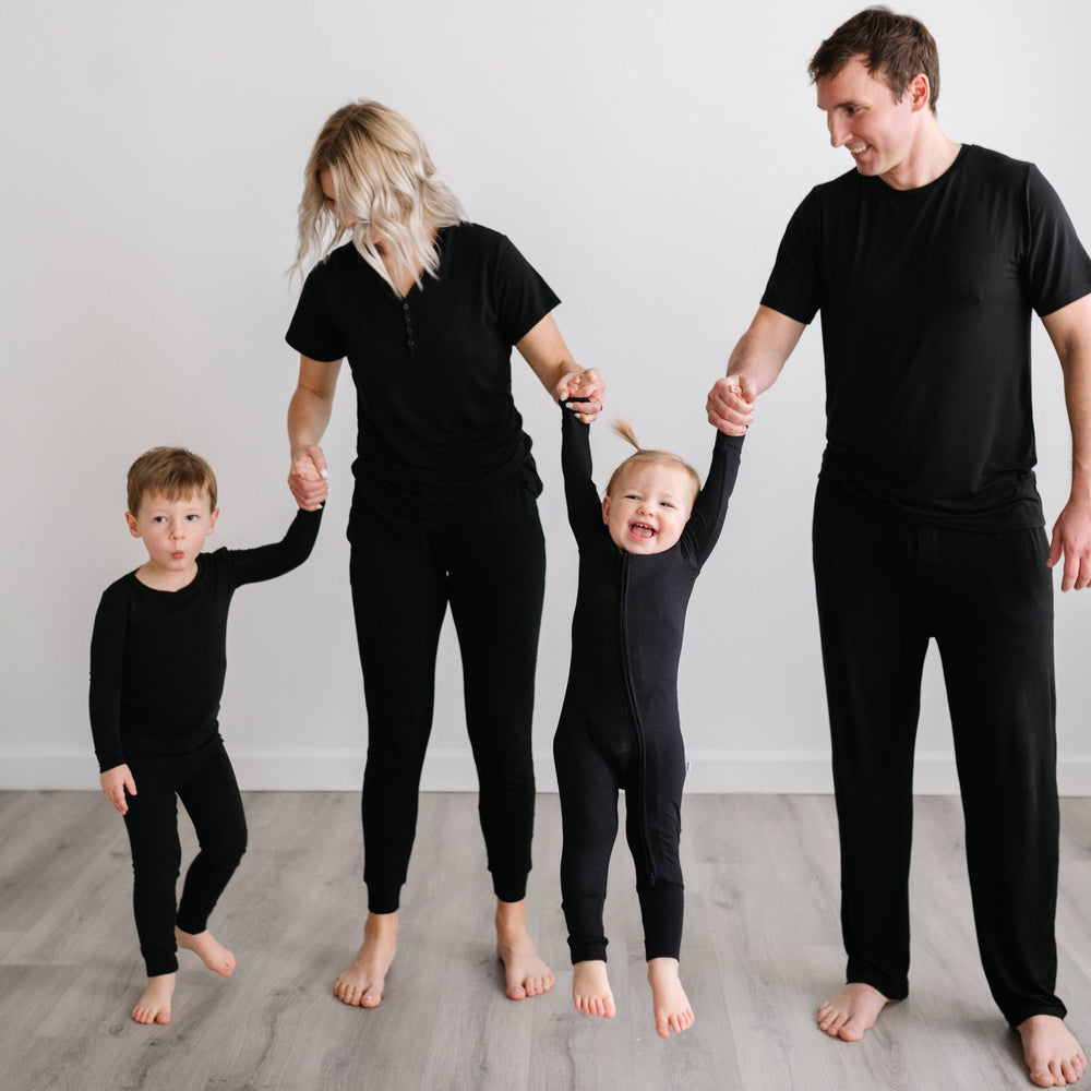 Click to see full screen - Image of family of 4 all holding hands. They are all wearing matching solid black pajamas. The mom and dad are both shown wearing solid black short sleeve pj tops with matching solid black pj pants, the son is shown wearing a long sleeve pajama set, and t