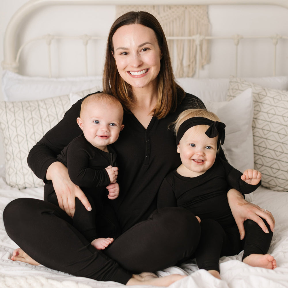 Mom, baby boy, and baby girl all wearing matching solid black pajamas. Mom is wearing solid black long sleeve pajama top and bottoms. Baby boy is wearing solid black zip up romper, while baby girl is wearing solid black pajama set with matching solid back bow headband. 
