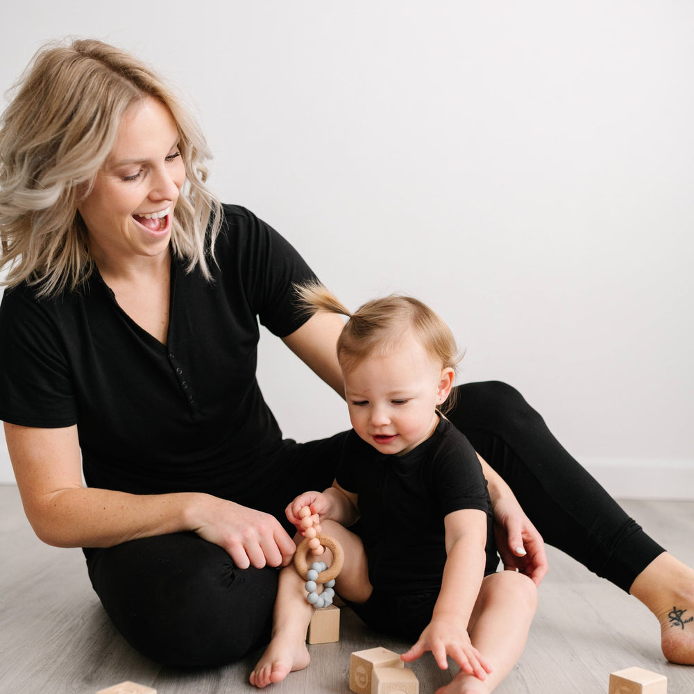 Image of mom and daughter playing with wooden building blocks. They are both shown wearing matching solid black pajamas. The mom is shown wearing a short sleeve pajama top with matching pj pants, and the toddler girl is shown wearing a short sleeve and sh