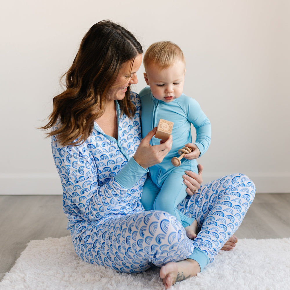 Image of mom holding her son. Mom is pictured wearing blue rainbow printed pajamas and baby boy is shown wearing sky blue zip up romper. 