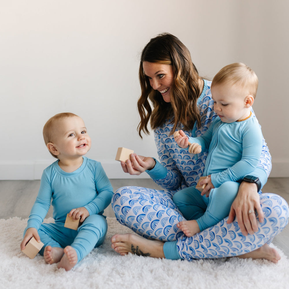 Image of mom and her two sons playing with building blocks. Mom is wearing blue rainbow printed pajamas and two boys are matching in their sky blue pajamas. 
