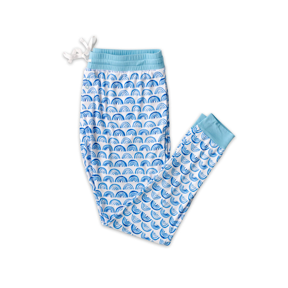 Flat lay photo of women's blue rainbow printed pajama pants. The pants are featured having pockets and a drawstring waist. This print sits on a white background with shades of blue rainbows and sky blue trim details.