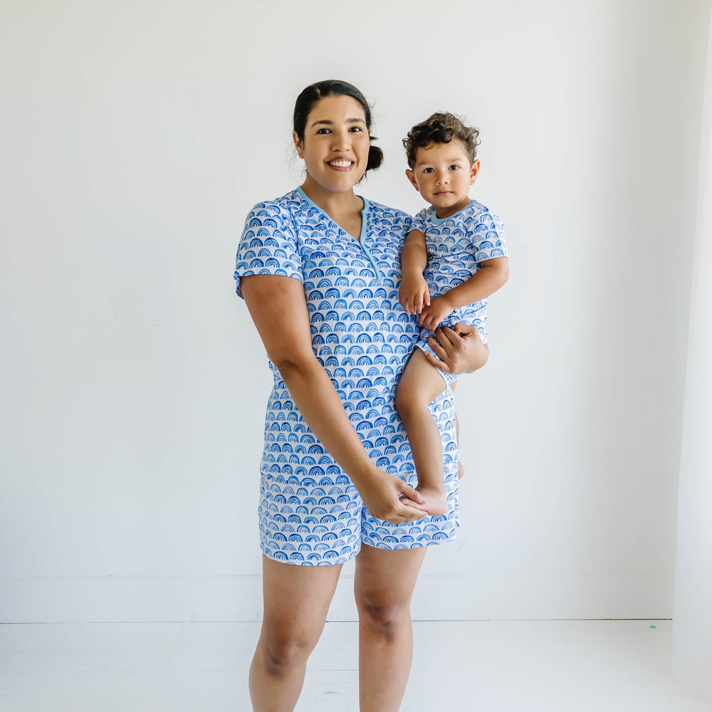 Click to see full screen - Image of mom holding her toddler son. They are both shown wearing matching blue rainbow printed short sleeve and shorts pajama sets. This print sits on a white background with shades of blue rainbows and sky blue trim details.