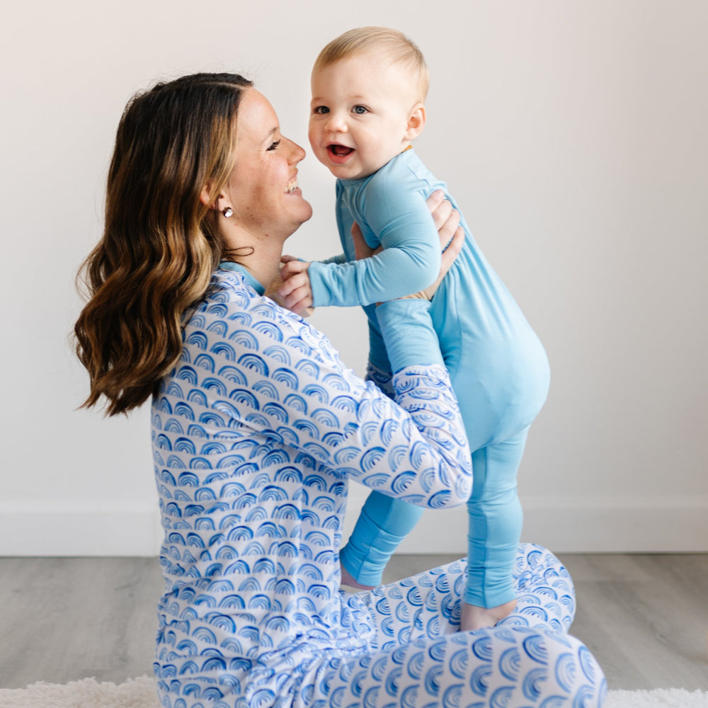 Image of mom holding her son. Mom is pictured wearing blue rainbow printed pajamas and baby boy is shown wearing sky blue zip up romper.