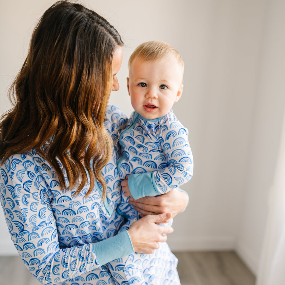 Image of mom and son wearing matching blue rainbow printed pajamas. This print sits on a white background with shades of blue rainbows and sky blue trim details.
