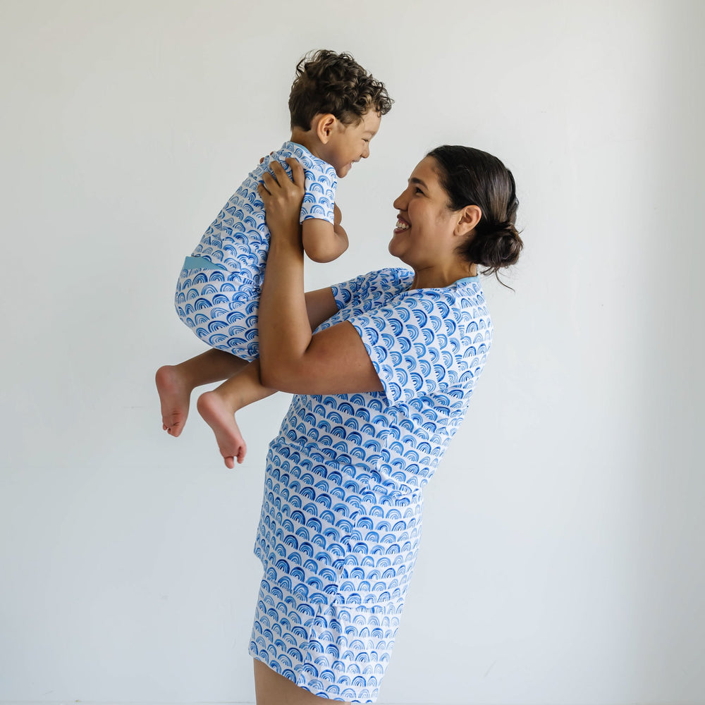 Image of mom holding her toddler son. They are both shown wearing matching blue rainbow printed short sleeve and shorts pajama sets. This print sits on a white background with shades of blue rainbows and sky blue trim details.