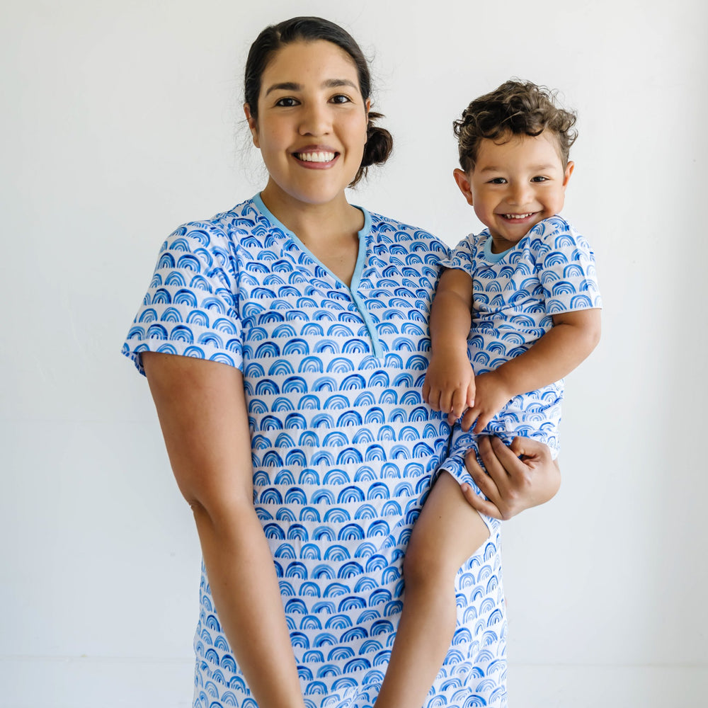 Image of mom holding her toddler son. They are both shown wearing matching blue rainbow printed short sleeve and shorts pajama sets. This print sits on a white background with shades of blue rainbows and sky blue trim details.