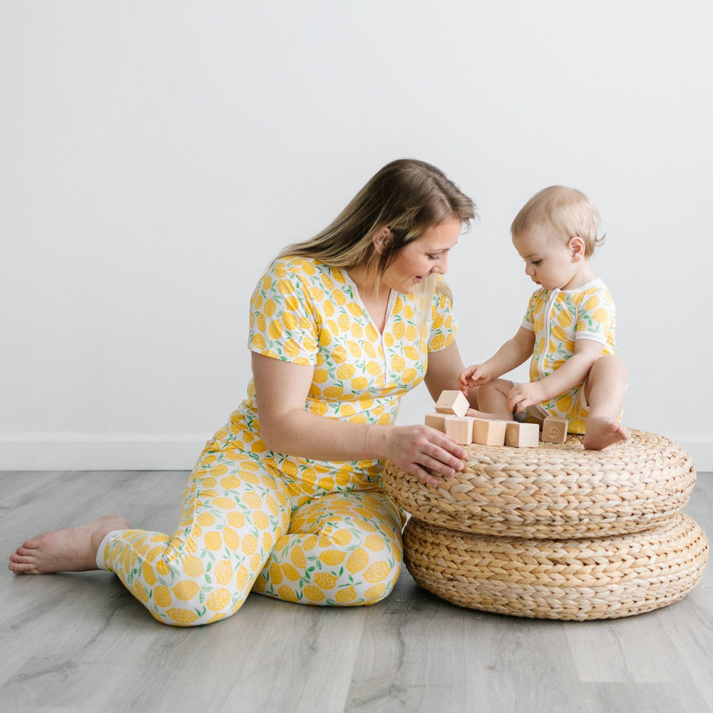 Image of mom and daughter playing with wooden building blocks. Both are shown wearing matching short sleeve pajama sets in Lemons print. This print features vibrant pops of yellow fruit with green accents that sit upon a white background with white trim.